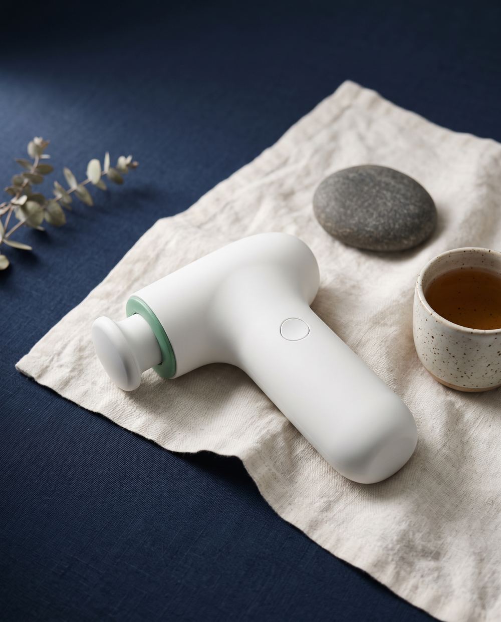 A white handheld massage device on linen beside a stone and a cup of tea